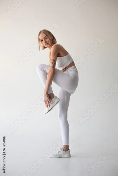 Fototapeta Beautiful blonde girl posing on a white background in white leggings and a white top