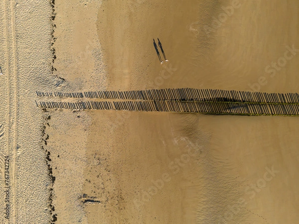 Fototapeta Bird's eye view of the sandy shore, long shadows on the sand from the wooden breakwater posts
