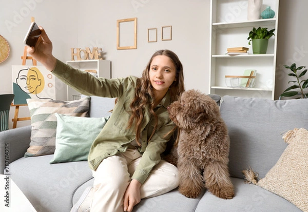 Fototapeta Young woman with cute poodle taking selfie on sofa at home