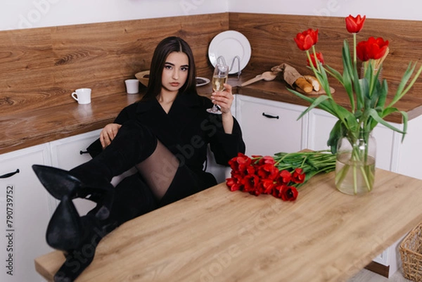 Fototapeta Woman poses with vibrant bouquets of red tulips in vase in comfortable kitchen hold glass of champagne, fashion outfit. Spring women`s day, 8th of March. 
