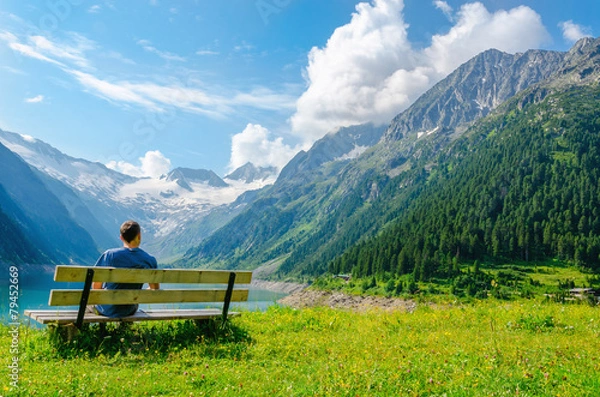 Fototapeta A young man sits on bench beside an azure mountain lake