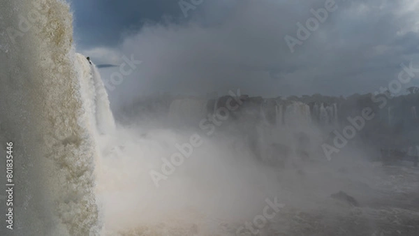 Fototapeta The powerful waterfall is shrouded in fog. Clouds in the blue sky. In the foreground, the flow of water is in close-up. Splashes, drops, and an openwork texture are visible. Iguazu Falls. Brazil