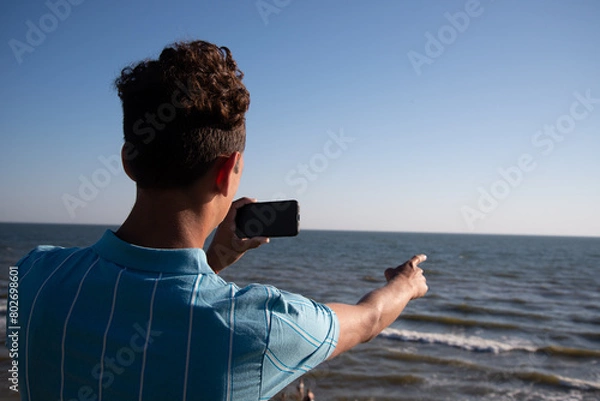 Fototapeta A young attractive guy is posing while relaxing on the sea and taking a photo with his phone.