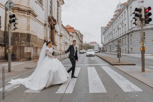 Fototapeta A bride and groom are walking across a street. The bride is wearing a white dress and the groom is wearing a suit. The couple is holding hands and walking together