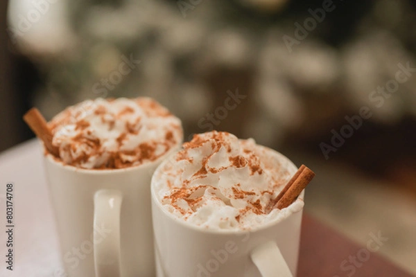 Fototapeta Close up of hot chocolate in a white ceramic mug with whipped cream and cinnamon powder & cinnamon sticks against a Christmas tree scene