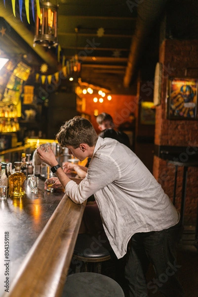 Fototapeta Drunk guy holds empty glass in hand and relax in patriotic nightclub. Visitor drinks vodka in club with subdued lights and brick walls