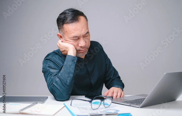 Fototapeta Sleepy businessman, an office manager, sits at his desk, working on his laptop, striving to stay productive despite his fatigue, highlighting relentless demands of his business responsibilities.