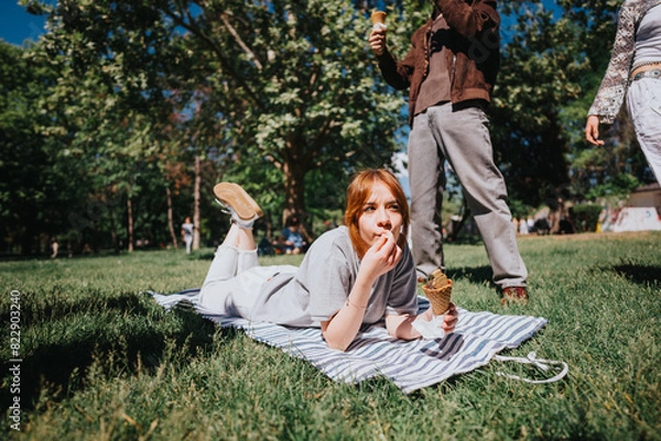 Fototapeta A relaxed young woman eating ice cream while lying on a blanket in a bustling park surrounded by friends on a sunny day.