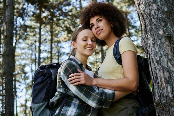 Fototapeta Two young women, a lesbian couple, stand together in a forest, embracing. They are both wearing backpacks and enjoying the wilderness.