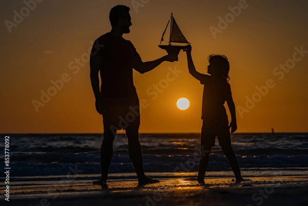 Fototapeta Father and son playing with toy sailboat, toy ship yacht on sea beach at summer sunrise. Sunset silhouette of father and son dreaming on cruise. Dreaming to travel with kid son. Fathers day.