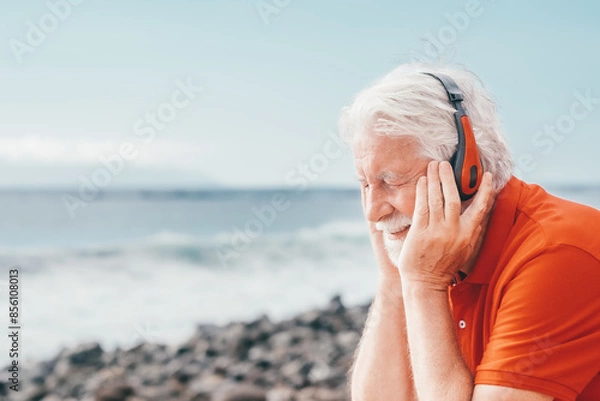 Fototapeta Smiling bearded senior man sitting in outdoors at the beach listening music by earphones, elderly male closed eyes enjoying  vacation, free time and retirement