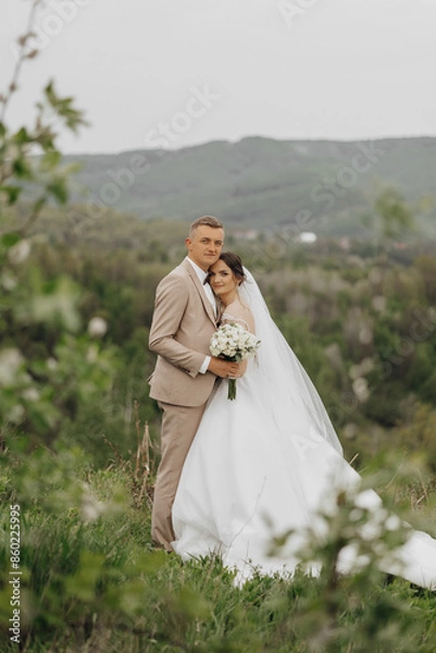 Fototapeta A bride and groom are standing on a grassy hillside, posing for a picture. The bride is wearing a white dress and the groom is wearing a suit. They are holding a bouquet and a bouquet of flowers