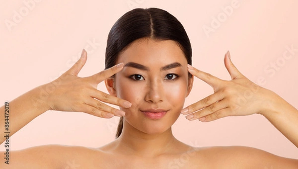 Fototapeta Asian young woman with long black hair, gently applying eye cream to her skin. She is looking directly at the camera with a smile and her hands are framing her face. The background is a soft pink.