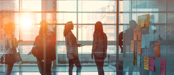 Fototapeta Group of silhouetted people engaged in discussion near a glass wall with sticky notes, bathed in the golden glow of sunset.