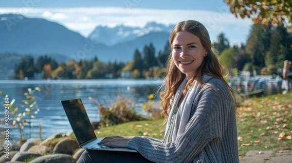 Fototapeta A young woman smiles as she sits on the grass beside a beautiful lake, working on her laptop, surrounded by fall foliage and mountains under a clear blue sky.